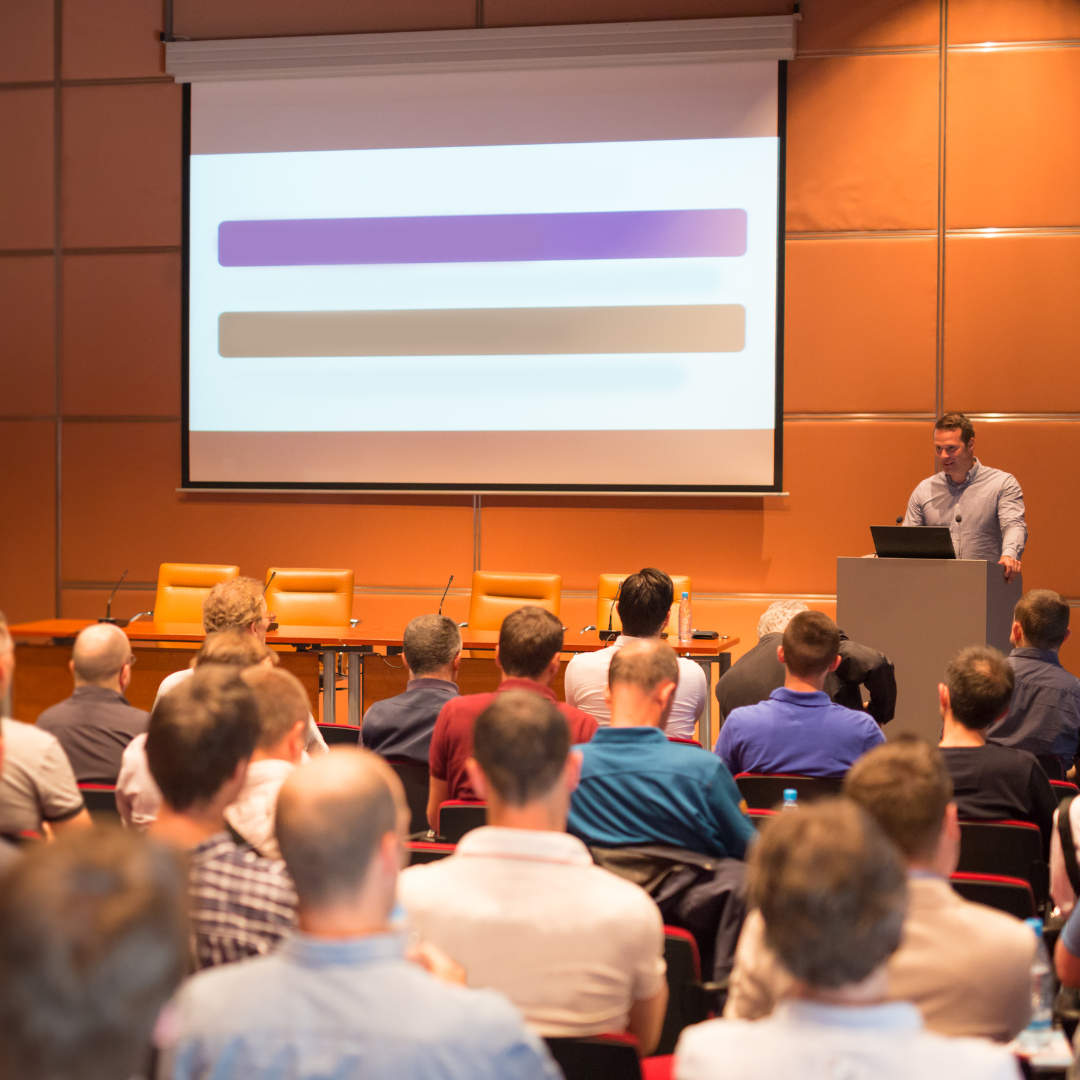 A speaker presenting to a seated audience in a conference room, with a large projection screen in the background.