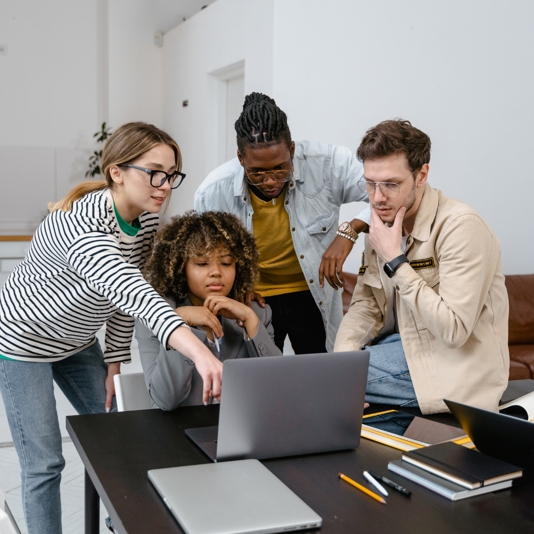 A diverse group of colleagues collaborating around a laptop in a modern office setting, with notebooks and pens on the table.