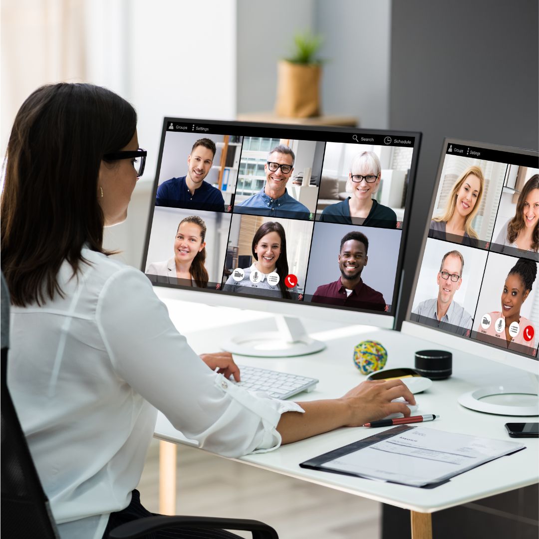 Woman working at a desk attending a virtual meeting on two computer screens, showing multiple participants on a video call.