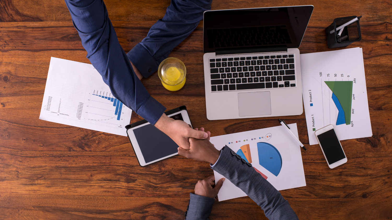 Two professionals shaking hands over a table with a laptop, tablet, phone, and financial charts, symbolizing collaboration.