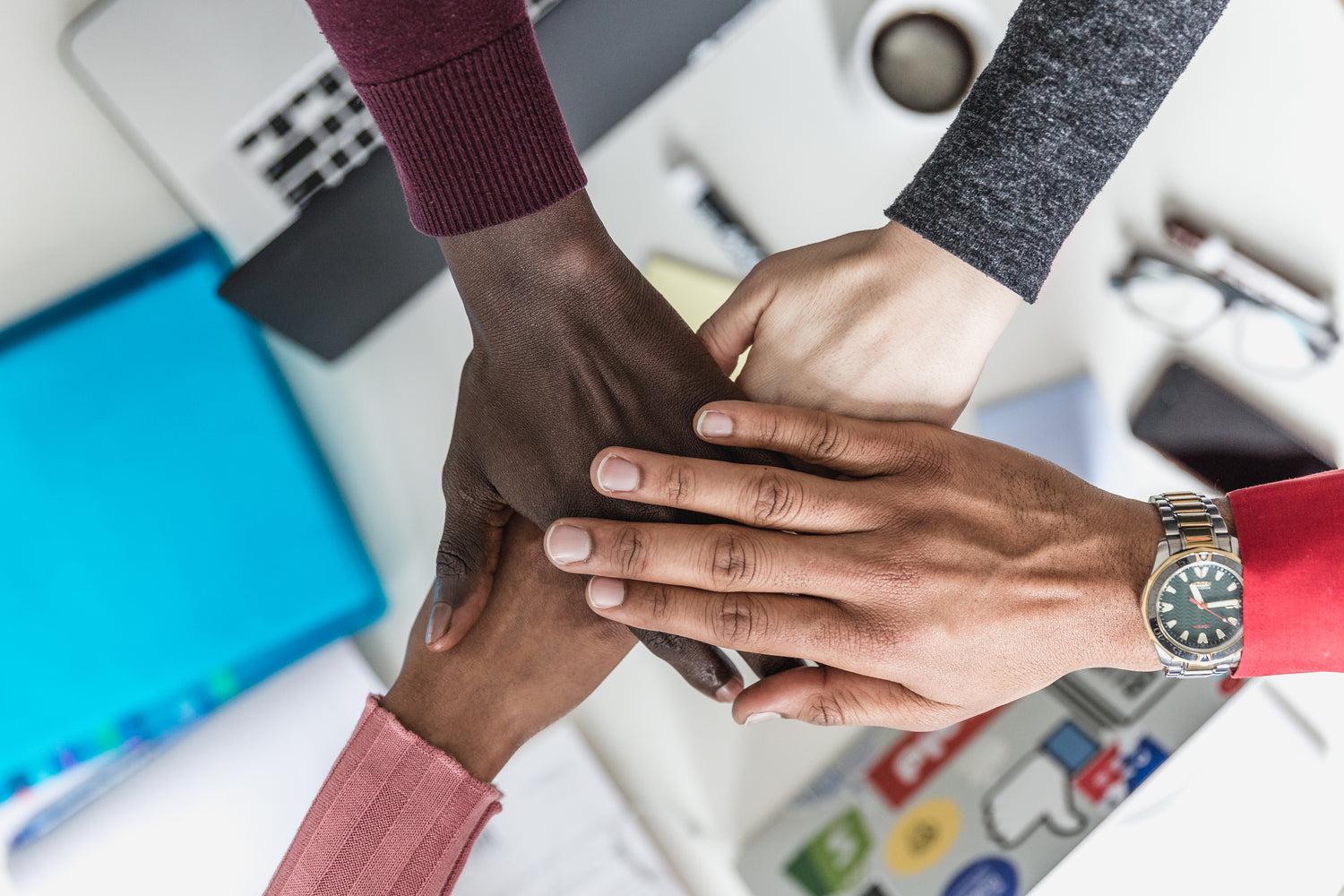 Close-up of a diverse group of hands stacked together in a teamwork gesture, placed over an office desk with various items.