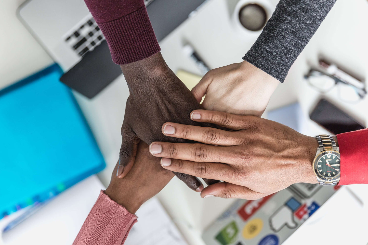 Close-up of a diverse group of hands stacked together in a teamwork gesture, placed over an office desk with various items.