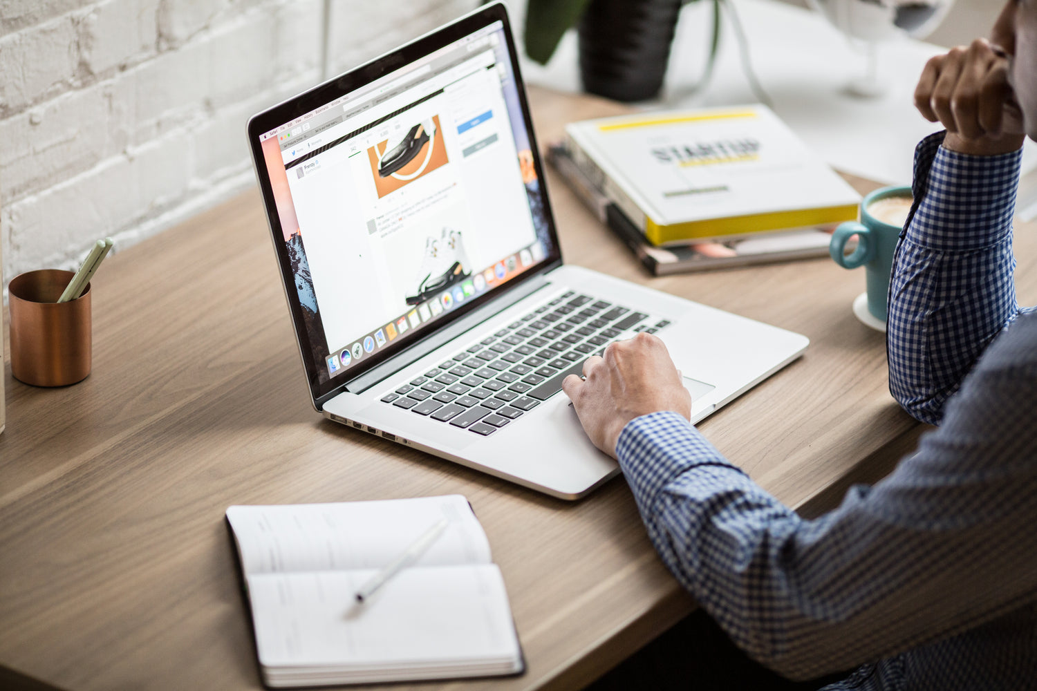 Person at a desk using a laptop displaying an online store, with a notebook, pen, coffee cup, and books nearby.