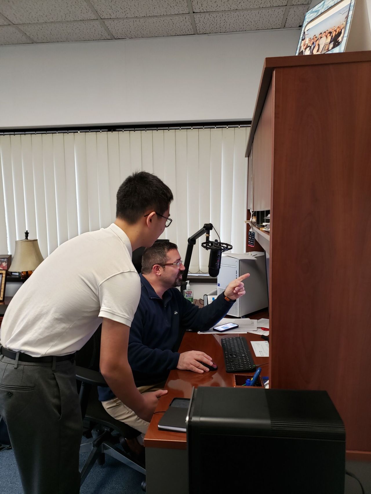 Two iemployees in an office, one sitting at a desk with a computer, the other pointing at the screen.