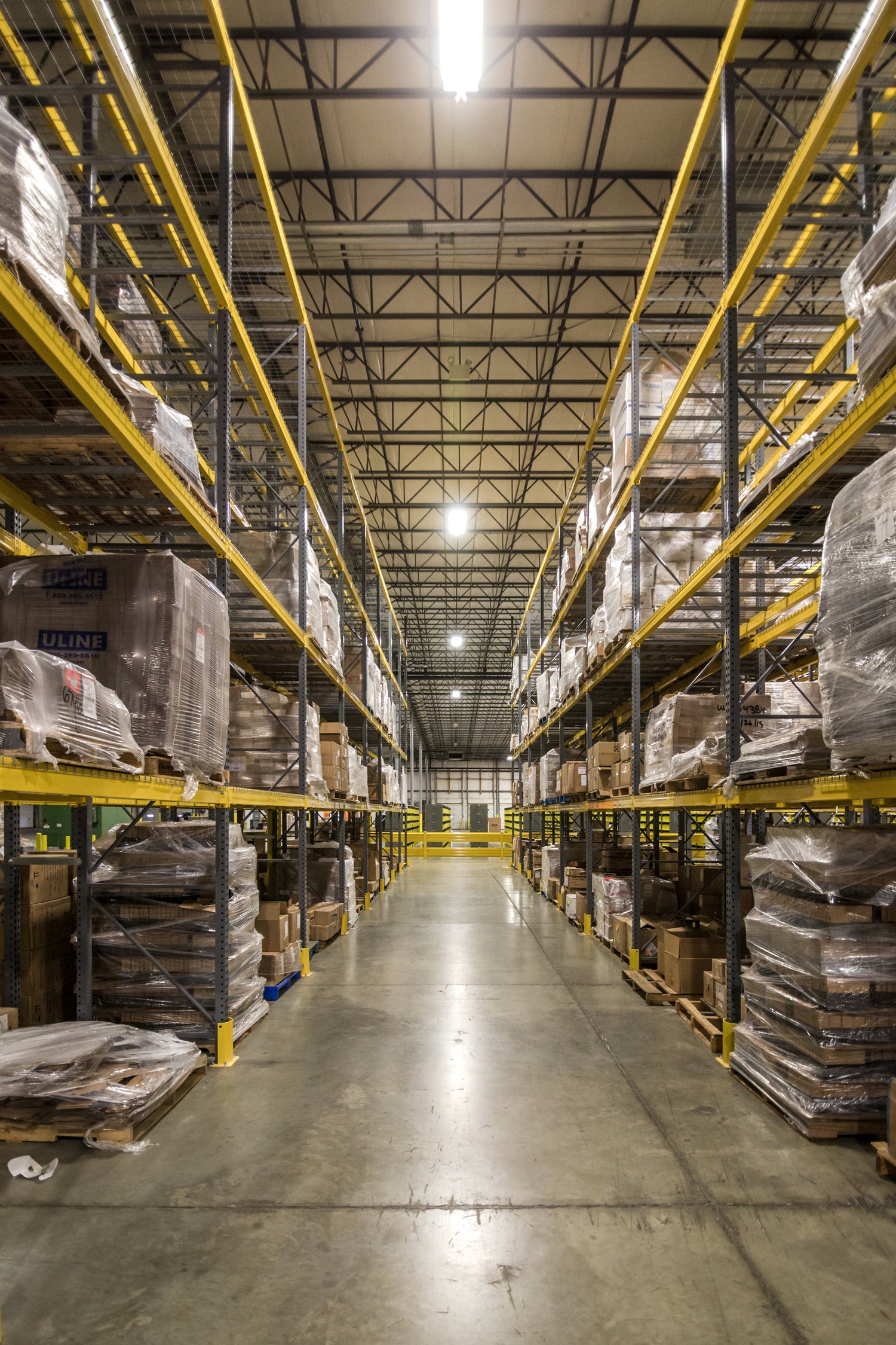Warehouse interior with tall yellow shelving racks filled with organized pallets and boxes, illuminated by overhead lights.