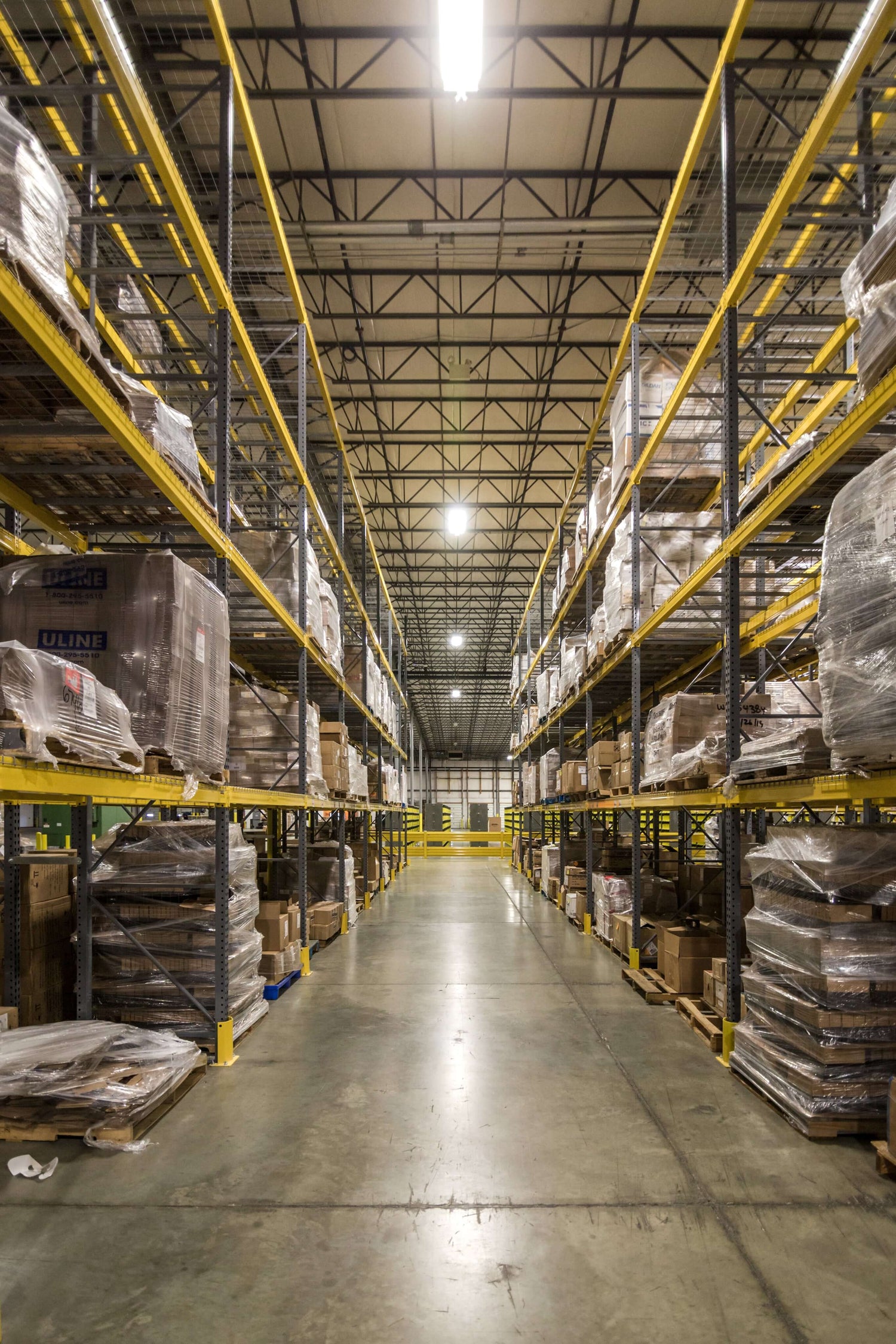 Warehouse interior with tall yellow shelving racks filled with organized pallets and boxes, illuminated by overhead lights.