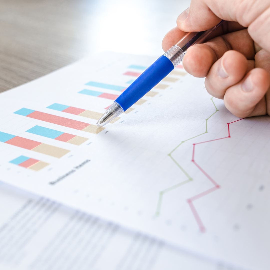 Close-up of a hand holding a pen, analyzing colorful bar and line graphs on a report at an Aptimized workspace.